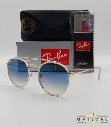 Lunettes de soleil Ray-Ban Round Double Bridge RB3647N dorées avec cerclages en acétate transparent et verres dégradés bleus, étui noir, boîte grise et livret de garantie chez Optical Gallery.