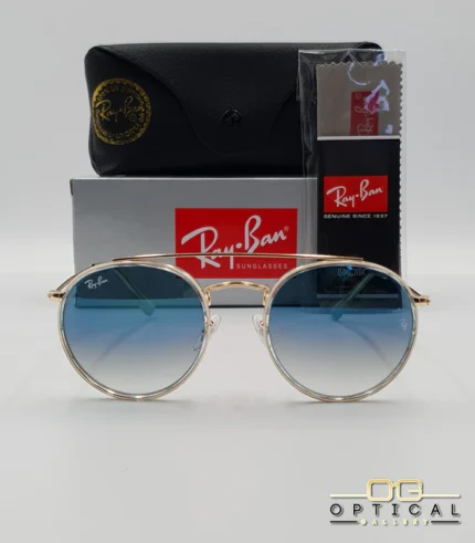Lunettes de soleil Ray-Ban Round Double Bridge RB3647N dorées avec cerclages en acétate transparent et verres dégradés bleus, étui noir, boîte grise et livret de garantie chez Optical Gallery.