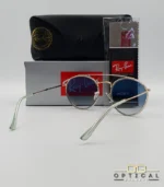 Lunettes de soleil Ray-Ban Round Double Bridge RB3647N dorées avec cerclages en acétate transparent et verres dégradés bleus, étui noir, boîte grise et livret de garantie chez Optical Gallery.