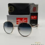 Lunettes de soleil Ray-Ban Round Double Bridge RB3647N dorées avec cerclages en acétate transparent et verres dégradés bleus, étui noir, boîte grise et livret de garantie chez Optical Gallery.