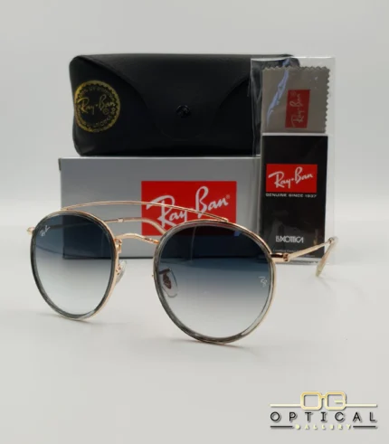 Lunettes de soleil Ray-Ban Round Double Bridge RB3647N dorées avec cerclages en acétate transparent et verres dégradés bleus, étui noir, boîte grise et livret de garantie chez Optical Gallery.