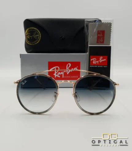 Lunettes de soleil Ray-Ban Round Double Bridge RB3647N dorées avec cerclages en acétate transparent et verres dégradés bleus, étui noir, boîte grise et livret de garantie chez Optical Gallery.