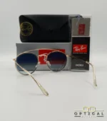 Lunettes de soleil Ray-Ban Round Double Bridge RB3647N dorées avec cerclages en acétate transparent et verres dégradés bleus, étui noir, boîte grise et livret de garantie chez Optical Gallery.