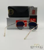 Lunettes de soleil Ray-Ban Round Double Bridge RB3647N dorées avec cerclages en acétate transparent et verres dégradés bleus, étui noir, boîte grise et livret de garantie chez Optical Gallery.