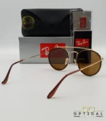 Lunettes de soleil Ray-Ban Round Double Bridge RB3647N dorées avec cerclages en acétate marron et verres marrons B-15, étui noir, boîte grise et livret de garantie chez Optical Gallery.