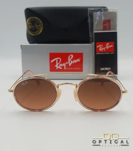Lunettes de soleil Ray-Ban Round Double Bridge RB3847N dorées avec cerclages en acétate rose transparent et verres dégradés marrons, étui noir, boîte grise et livret de garantie chez Optical Gallery.