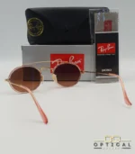 Lunettes de soleil Ray-Ban Round Double Bridge RB3847N dorées avec cerclages en acétate rose transparent et verres dégradés marrons, étui noir, boîte grise et livret de garantie chez Optical Gallery.