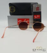 Lunettes de soleil Ray-Ban Round Double Bridge RB3847N dorées avec cerclages en acétate rose transparent et verres dégradés marrons, étui noir, boîte grise et livret de garantie chez Optical Gallery.